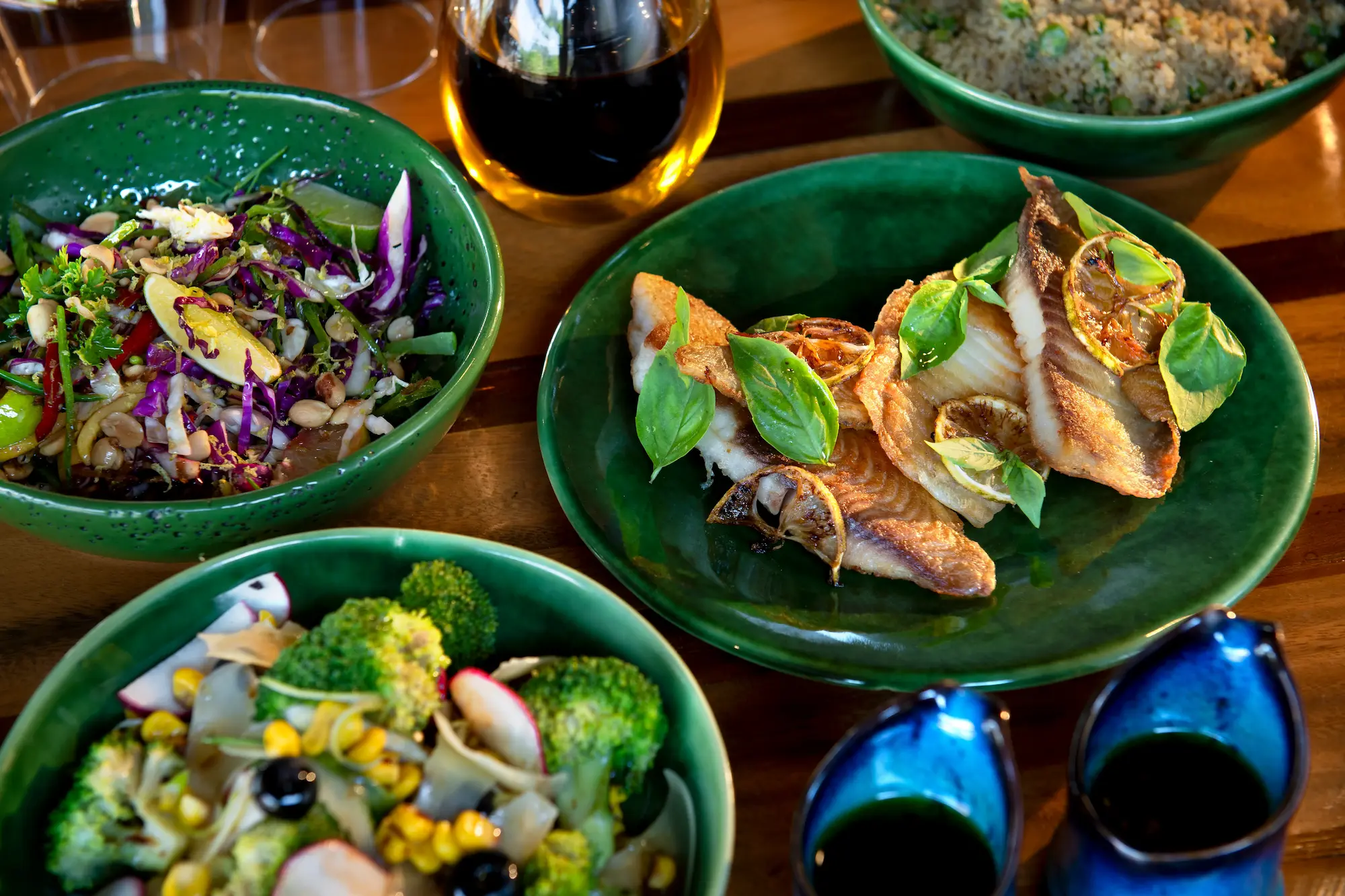 A delicious meal served at Atzaro Okavango, featuring a variety of dishes on green plates. The meal includes a salad with mixed greens, a salad with broccoli and corn, and a plate of fried fish with lemon and herbs.