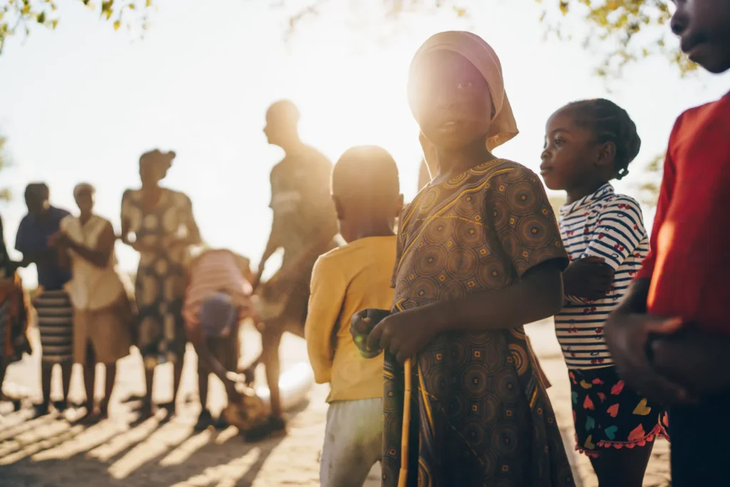 a small child faces the camera. In the background adults and several other children are gathered together wearing bright colours