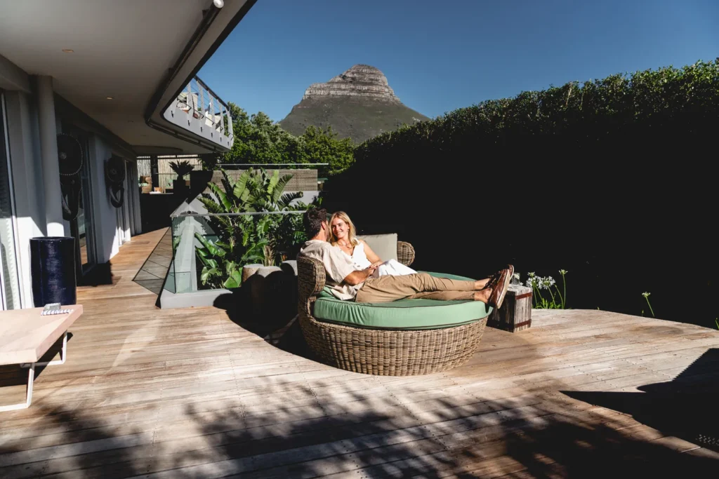 A couple recline on a round sun lounger that's placed on a wooden deck in the sunshine. In the background, a rugged mountain peak is visible.