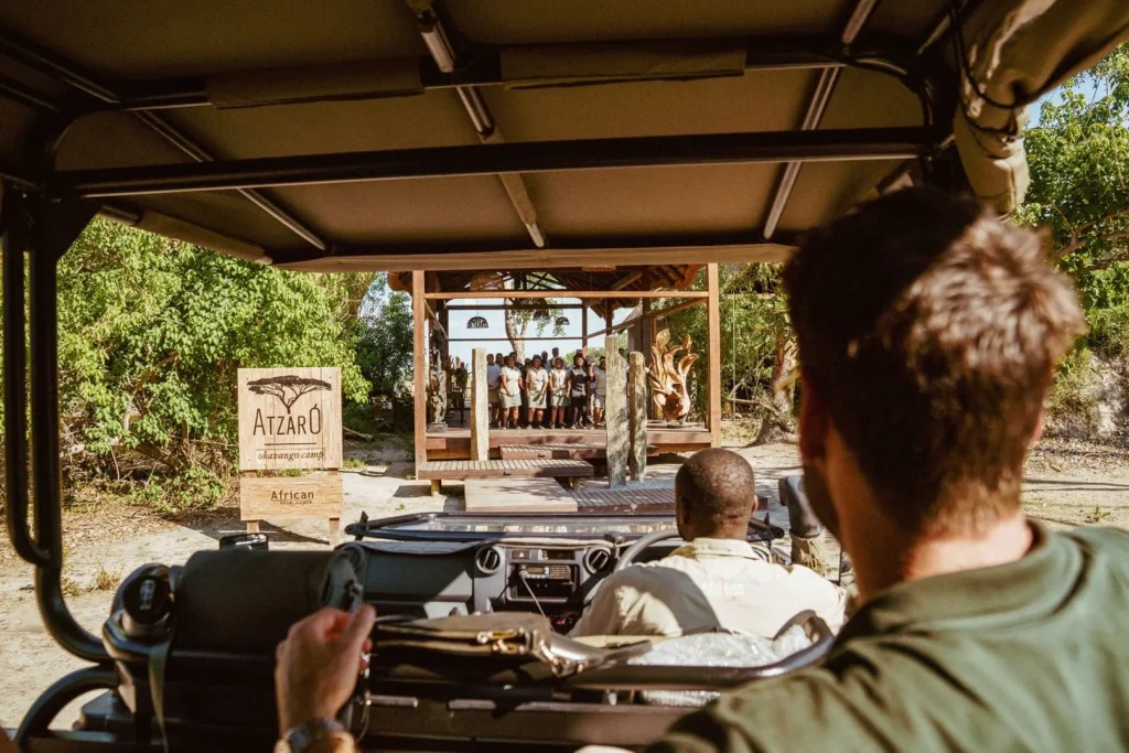 Two men in a game viewing vehicle approach a wooden platform where a group of people are dancing, waiting to welcome them into the safari lodge