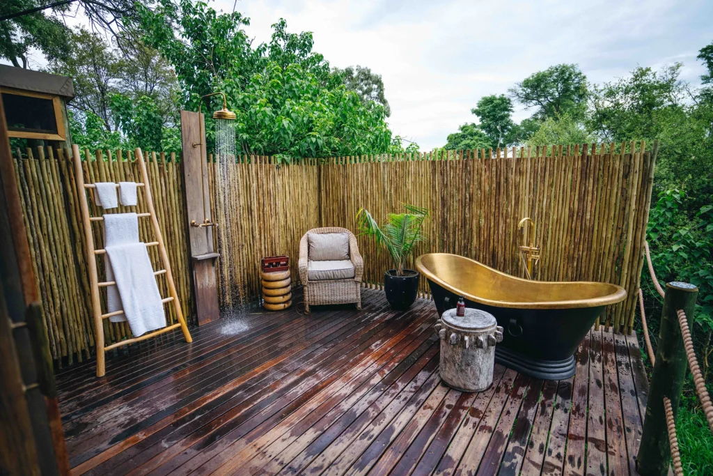 A luxurious private outdoor bathroom at Atzaró Okavango, featuring a brass bathtub and open-air shower on a spacious wooden terrace overlooking the wilderness.