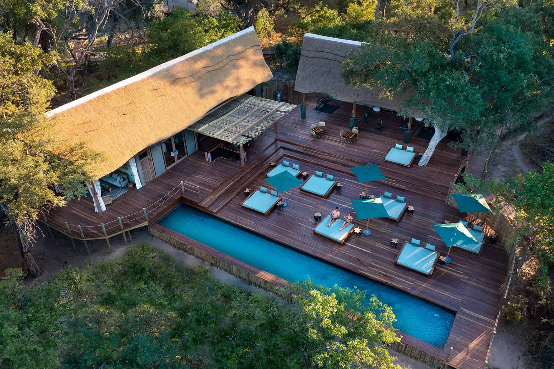 A stunning aerial view of Atzaro Okavango, showcasing the main lodge building with a thatched roof, a swimming pool, and a spacious outdoor deck with sun loungers. The image highlights the luxurious and tranquil setting of the lodge.