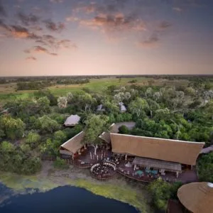 Aerial drone view of Atzaró Okavango Camp showing thatched luxury suites and main lodge nestled in lush greenery along a lagoon in the Okavango Delta.