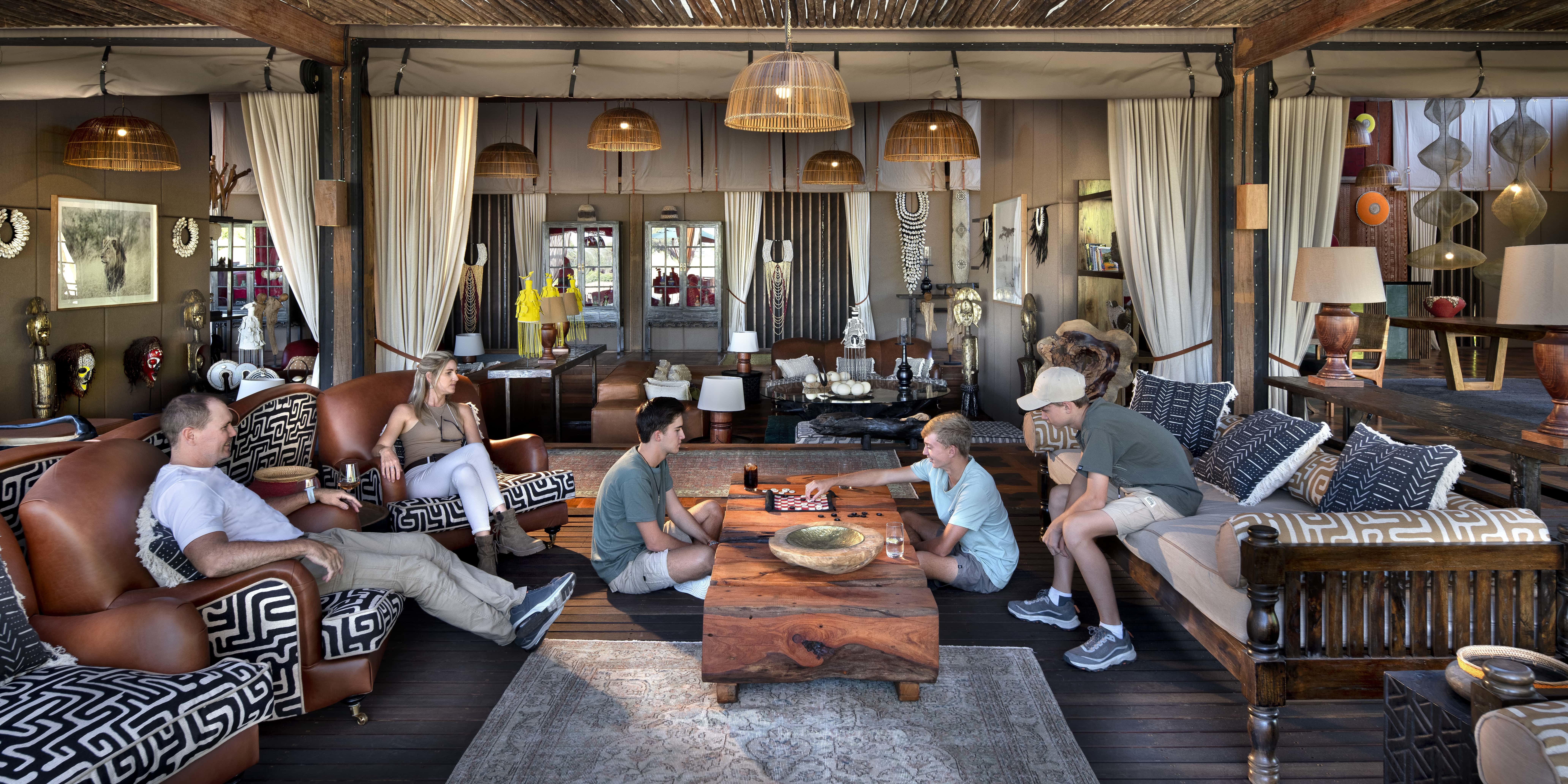 A family relaxing and playing board games in the luxury common area of Atzaró Okavango Camp, featuring elegant African decor and a safe, spacious lounge.