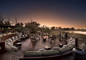 Guests gathering around a central fire pit on the sunken lounge deck at Atzaró Okavango Camp during a vibrant sunset.