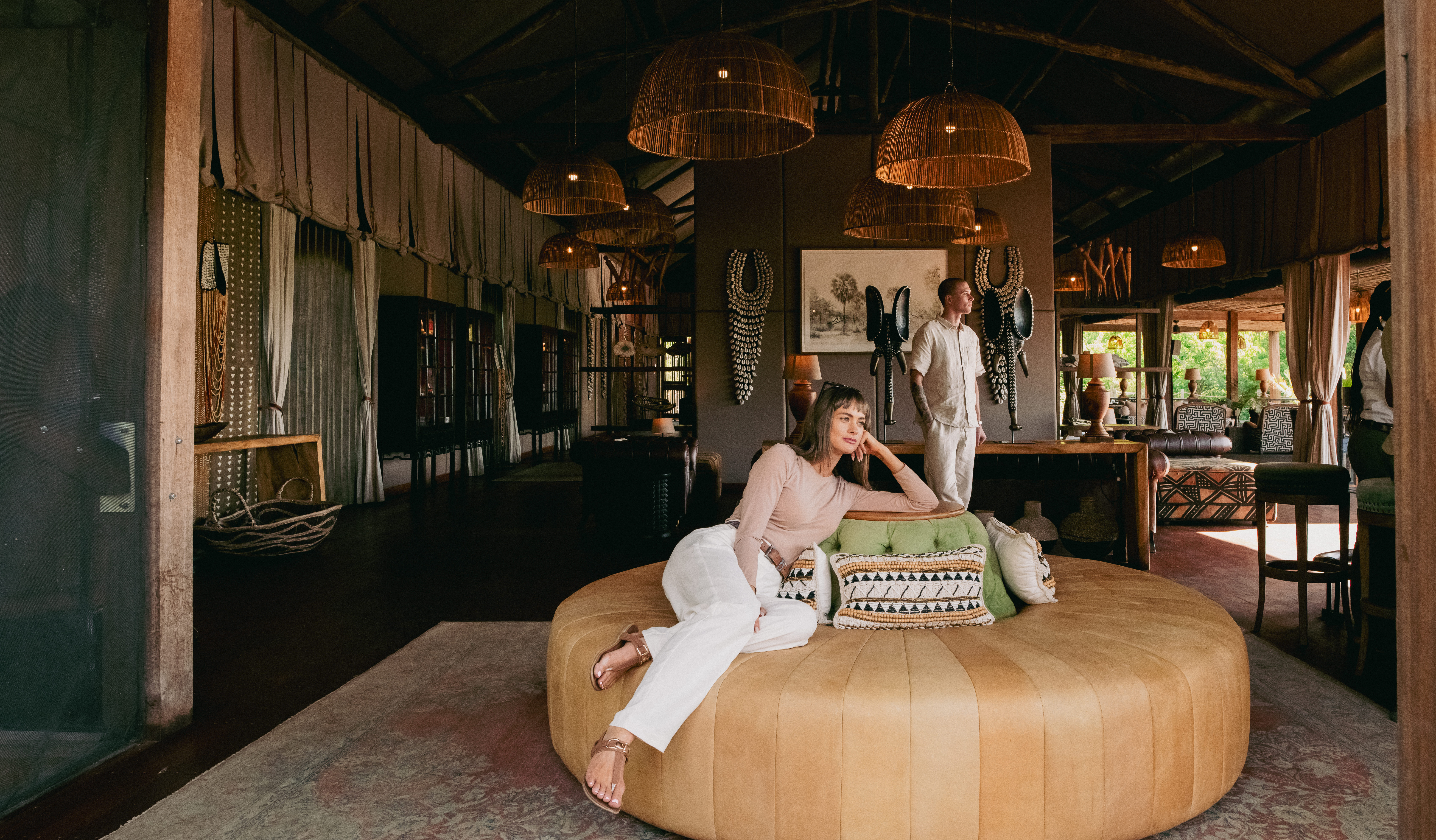 A woman sitting on a large round leather daybed while a man stands behind her in the luxurious open-air main lounge of Atzaró Okavango Camp, surrounded by woven pendant lights and African decor.
