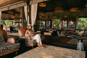 A female guest wearing a straw sun hat sits comfortably in a patterned leather armchair inside the open-air luxury safari lounge at Atzaró Okavango Camp.