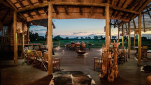Open-air Ancestral Boma with tall wooden posts, a thatched roof and safari chairs around a central fire bowl, a cowhide rug in the foreground and green Delta plains glowing beyond in the evening light.