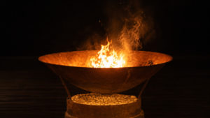 Copper-brown fire bowl blazing with bright orange flames over glowing stones against the black night, the central fire where guests gather for a boma dinner at Atzaró Okavango.