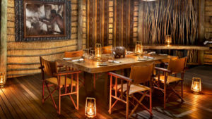 Rustic dining table set with glass goblets and lit lanterns, surrounded by leather and wood safari chairs, timber walls, carved wooden sculptures and a framed black and white portrait, a warm boma dinner setting.
