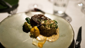 Plated seared beef fillet with creamy sauce, glossy green vegetables and bright yellow squash on a pale sage-green plate, a seasonal farm-to-table dish from the in-house chefs at Atzaró Okavango.