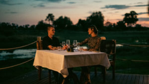 Couple sharing a romantic dinner at a table with a cream tablecloth and wine glasses on a deck above the lagoon, with dark silhouetted trees and a teal and soft pink dusk sky behind them.