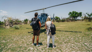 Couple with dark backpacks walking across a green airstrip toward a waiting dark blue helicopter, with scattered trees and a bright blue sky ahead, the short private flight from Maun Airport into the Okavango Delta. 