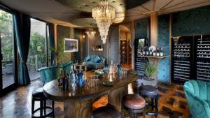 Elegant Wine Cellar lounge with a crystal chandelier, a long dark wood table set with glasses and blue bottles, teal velvet armchairs, glass wine fridges and tall windows, made for private tastings and sommelier pairings.